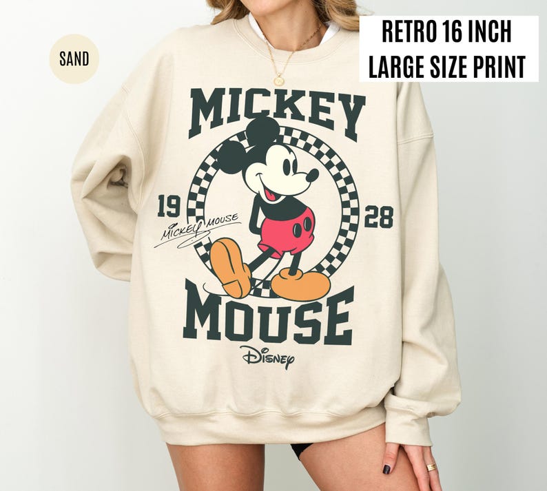 Może przedstawiać: Beżowa bluza z dużym nadrukiem Myszki Miki. Projekt zawiera tekst "Mickey Mouse" i "Disney" z rokiem "1928". Bluza ma nadruk w stylu retro o wielkości 40,6 cm.