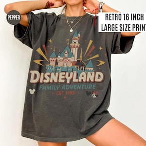 Puede incluir: Camiseta gris oscuro con un gr&aacute;fico de Disneyland. El gr&aacute;fico presenta un castillo, la palabra "Disneyland" y el texto "Family Adventure Est. 1955". La impresi&oacute;n mide 40.6 cm de ancho. La camiseta tiene mangas cortas.