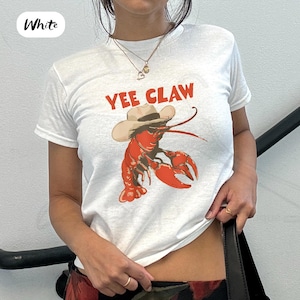 May include: White t-shirt featuring a red lobster wearing a cowboy hat with the text "Yee Claw" in red. The lobster graphic is a playful design. The shirt is a casual, short-sleeved style.