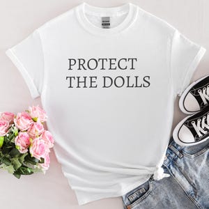 Puede incluir: Camiseta blanca con el texto "PROTECT THE DOLLS" impreso en negro.