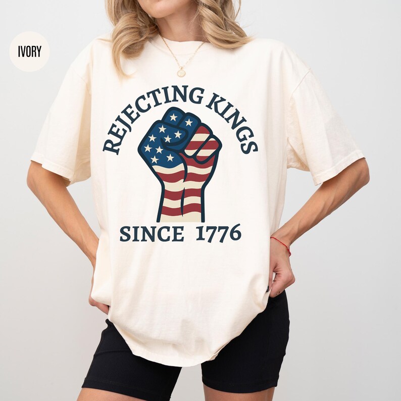 Puede incluir: Camiseta de color marfil con el texto "REJECTING KINGS SINCE 1776" y un pu&ntilde;o americano con los colores de la bandera estadounidense. La camiseta tiene mangas cortas y cuello redondo.