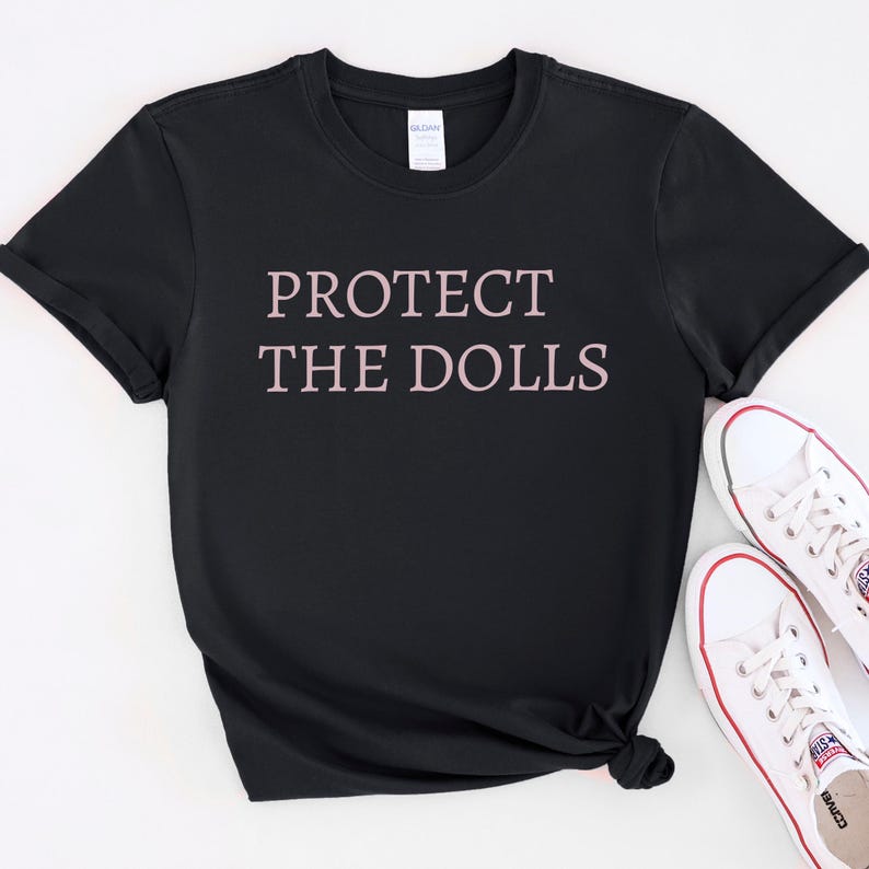 Puede incluir: Camiseta negra con el texto "PROTECT THE DOLLS" impreso en rosa en la parte delantera.
