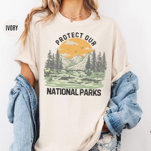 May include: Ivory t-shirt with a vintage-style graphic of a mountain landscape and the text "Protect Our National Parks".