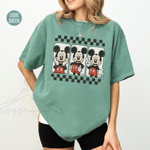 Puede incluir: Camiseta verde claro con un gr&aacute;fico de Mickey Mouse en tres poses diferentes. El gr&aacute;fico est&aacute; enmarcado por un patr&oacute;n de cuadros blancos y negros. La camiseta es de corte holgado.
