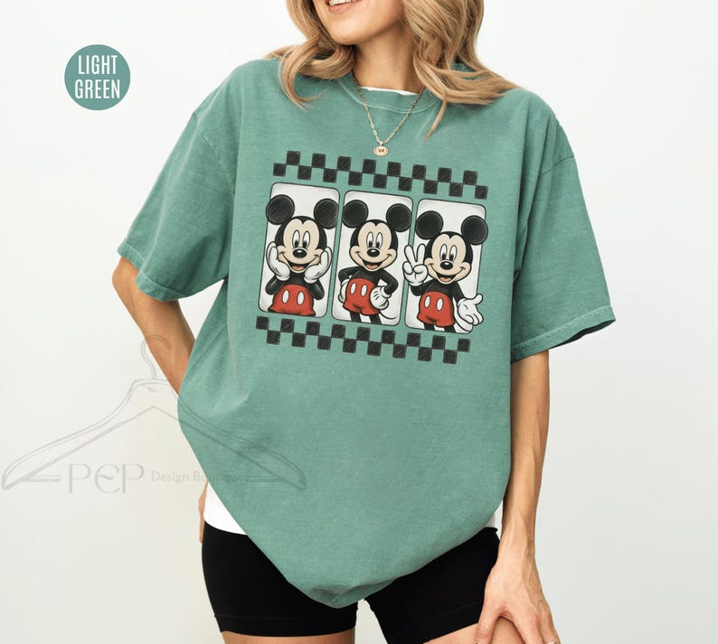 Puede incluir: Camiseta verde claro con un gr&aacute;fico de Mickey Mouse en tres poses diferentes. El gr&aacute;fico est&aacute; enmarcado por un patr&oacute;n de cuadros blancos y negros. La camiseta es de corte holgado.