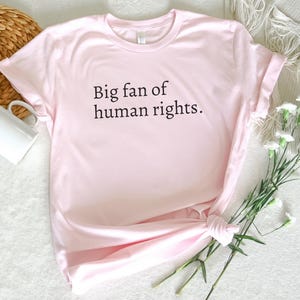 May include: A light pink t-shirt with the text "Big fan of human rights" in black. The shirt is knotted at the bottom and is next to a white mug and a bouquet of white flowers.