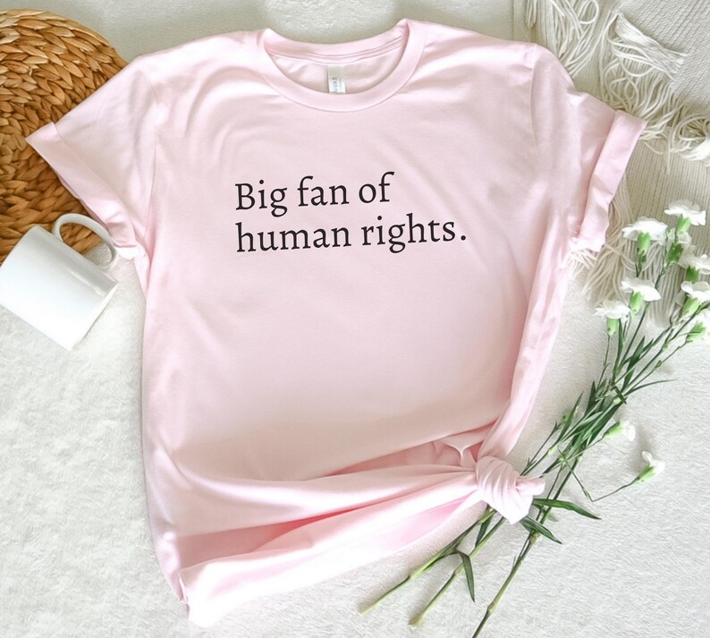May include: A light pink t-shirt with the text "Big fan of human rights" in black. The shirt is knotted at the bottom and is next to a white mug and a bouquet of white flowers.