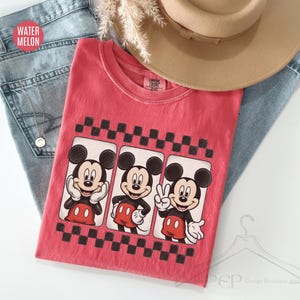 Puede incluir: Una camiseta roja con tres im&aacute;genes de Mickey Mouse en marcos en blanco y negro, sobre un patr&oacute;n de tablero de ajedrez en blanco y negro. La camiseta est&aacute; sobre unos vaqueros azules y un sombrero beige. El texto "WATER MELON" es visible.