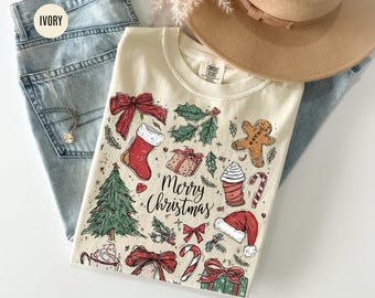 May include: Cream-coloured t-shirt featuring a "Merry Christmas" design with festive illustrations. The design includes a Christmas tree, stocking, gifts, gingerbread man, candy cane, and holly. The shirt is on blue jeans and a tan hat.