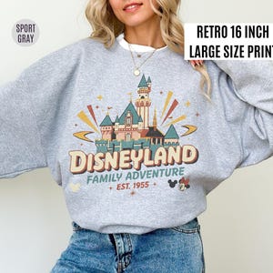 May include: A light gray sweatshirt with a retro Disneyland graphic. The graphic features a castle, the word "Disneyland", and the text "Family Adventure Est. 1955". The sweatshirt has a white collar and the text "Retro 16 inch Large Size Print".