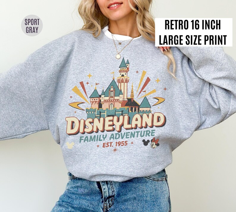 May include: A light gray sweatshirt with a retro Disneyland graphic. The graphic features a castle, the word "Disneyland", and the text "Family Adventure Est. 1955". The sweatshirt has a white collar and the text "Retro 16 inch Large Size Print".