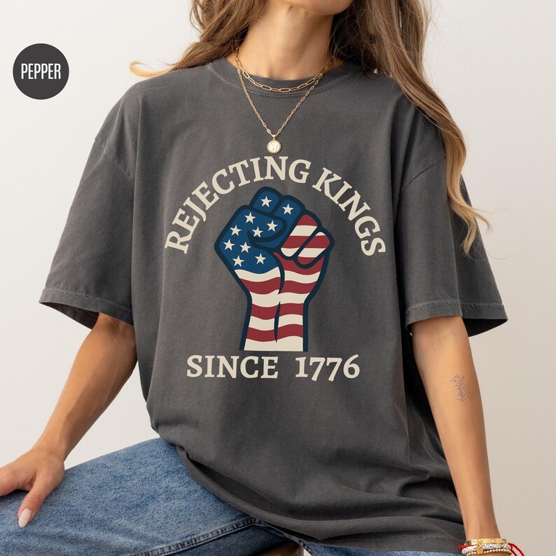 Puede incluir: Camiseta gris oscuro con el texto "Rejecting Kings Since 1776" que rodea un pu&ntilde;o con los colores de la bandera estadounidense. La camiseta tiene mangas cortas y un corte holgado.