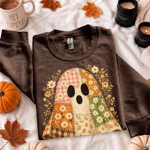 Op de afbeelding: Een donkerbruin sweatshirt met lange mouwen met een centrale afbeelding van een geest samengesteld uit verschillende patchwork-stofpatronen, waaronder bloemen-, gingham- en ruitprints, omringd door een decoratieve rand van kleine witte en gele bloemen.