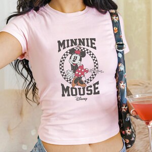 K&ouml;nnte beinhalten: Hellrosa T-Shirt mit Minnie Mouse-Grafik. Das Design zeigt die Worte "MINNIE MOUSE" in Schwarz, mit Minnie Mouse in der Mitte. Das Disney-Logo befindet sich unten. Das T-Shirt hat kurze &Auml;rmel und einen Rundhalsausschnitt.