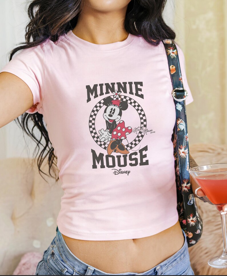 K&ouml;nnte beinhalten: Hellrosa T-Shirt mit Minnie Mouse-Grafik. Das Design zeigt die Worte "MINNIE MOUSE" in Schwarz, mit Minnie Mouse in der Mitte. Das Disney-Logo befindet sich unten. Das T-Shirt hat kurze &Auml;rmel und einen Rundhalsausschnitt.