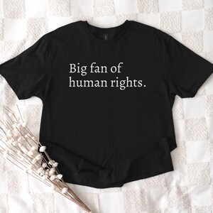 May include: Black t-shirt with the text "Big fan of human rights" in white. The shirt is displayed on a white patterned surface, with a bundle of white flowers in the lower left corner.