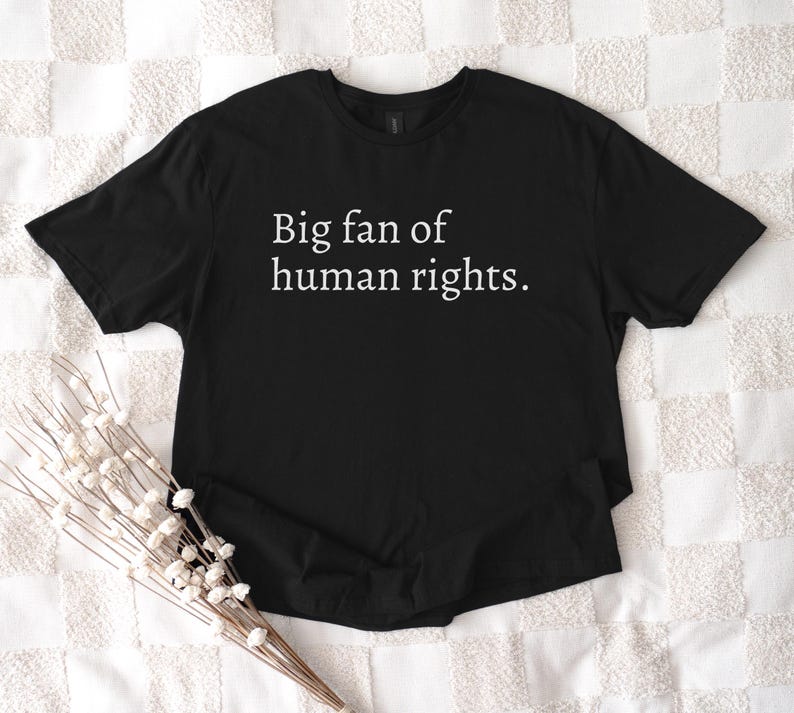 May include: Black t-shirt with the text "Big fan of human rights" in white. The shirt is displayed on a white patterned surface, with a bundle of white flowers in the lower left corner.