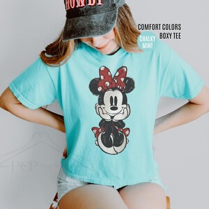 May include: A light blue, boxy tee shirt featuring a distressed graphic of Minnie Mouse with a red polka dot bow. The shirt has the words "Comfort Colors" and "Chalky Mint" printed on the upper right side.