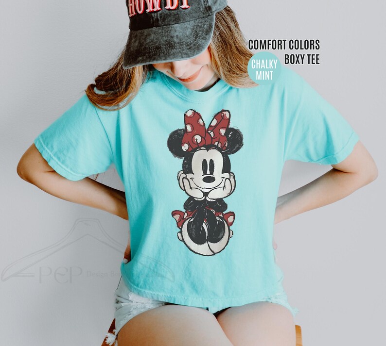 May include: A light blue, boxy tee shirt featuring a distressed graphic of Minnie Mouse with a red polka dot bow. The shirt has the words "Comfort Colors" and "Chalky Mint" printed on the upper right side.