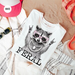 May include: White t-shirt with a raccoon graphic wearing pink sunglasses and a pearl necklace. The word "FERAL" is printed below the raccoon. A pink and white trucker hat, sunglasses, and coral sandals are also in the image.