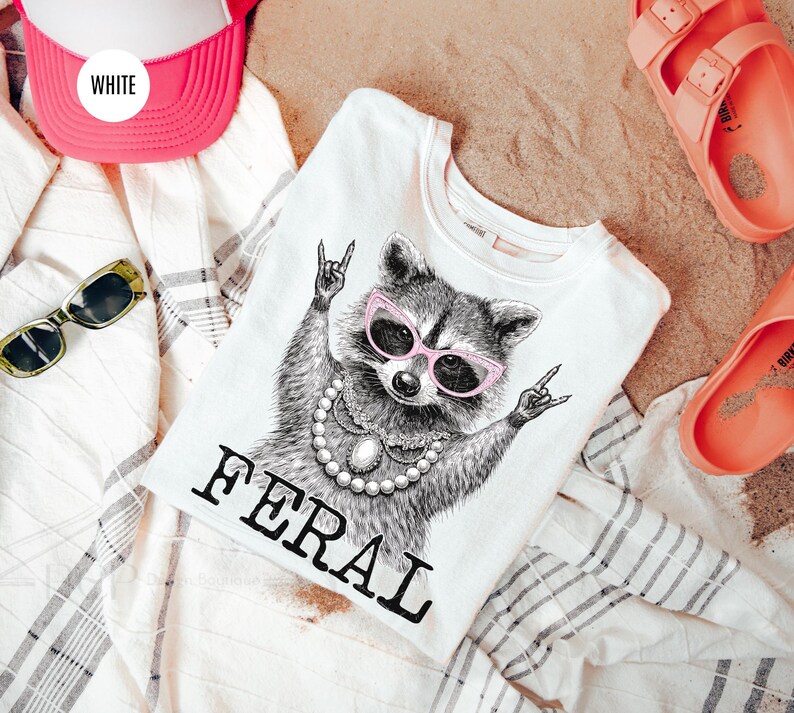 May include: White t-shirt with a raccoon graphic wearing pink sunglasses and a pearl necklace. The word "FERAL" is printed below the raccoon. A pink and white trucker hat, sunglasses, and coral sandals are also in the image.