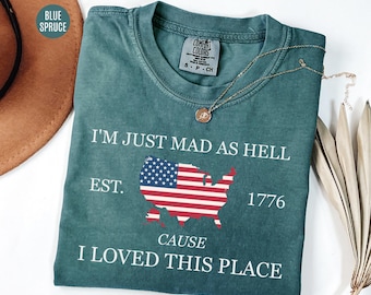 May include: Blue spruce-colored t-shirt with the text "I'M JUST MAD AS HELL CAUSE I LOVED THIS PLACE" and an American flag design. The shirt has a crew neck and short sleeves. A gold necklace is draped across the shirt.