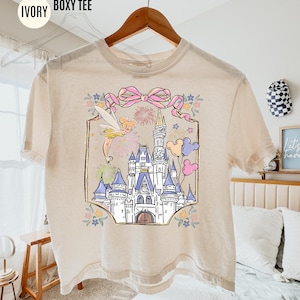 May include: Ivory boxy tee with a colorful graphic of a fairytale castle, fairy, and balloons. The graphic is framed with a pink bow and floral accents. The shirt has the words "COMFORT COLORS" and "IVORY BOXY TEE" printed on it.