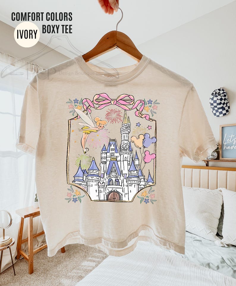 May include: Ivory boxy tee with a colorful graphic of a fairytale castle, fairy, and balloons. The graphic is framed with a pink bow and floral accents. The shirt has the words "COMFORT COLORS" and "IVORY BOXY TEE" printed on it.