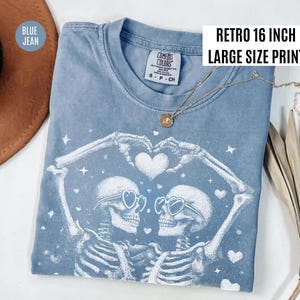 May include: Blue jean-colored t-shirt with a white graphic of two skeletons forming a heart with their arms. The skeletons wear heart-shaped sunglasses. The shirt has the text "Retro 16 inch Large Size Print". A gold necklace is draped on the shirt.