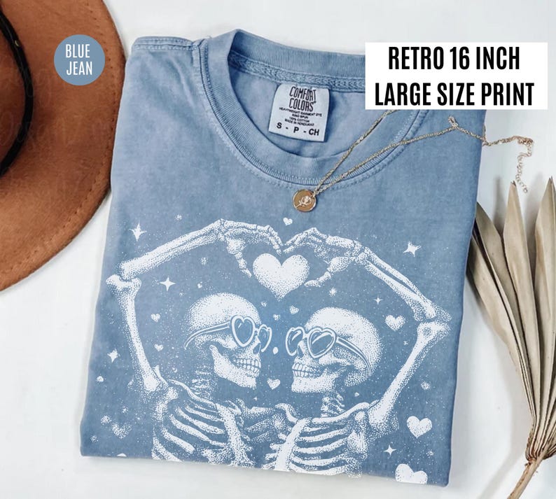 May include: Blue jean-colored t-shirt with a white graphic of two skeletons forming a heart with their arms. The skeletons wear heart-shaped sunglasses. The shirt has the text "Retro 16 inch Large Size Print". A gold necklace is draped on the shirt.