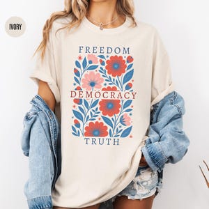 Op de afbeelding: Ivoor kleurig T-shirt met een bloemenontwerp met rode en roze bloemen, blauwe bladeren en de woorden "FREEDOM DEMOCRACY TRUTH" in het rood. Het shirt wordt gedragen met een spijkerjasje en een korte broek.