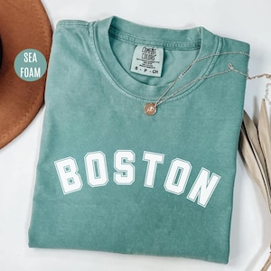 Op de afbeelding: Een tealblauw t-shirt met het woord "BOSTON" bedrukt in witte blokletters. Het shirt is gemaakt van een zachte, comfortabele stof en heeft een ontspannen pasvorm.