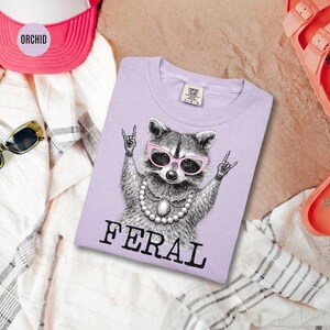 May include: A light purple t-shirt featuring a raccoon wearing pink sunglasses and a pearl necklace, with the word "FERAL" printed in black. The raccoon is making a rock and roll hand gesture. The shirt is surrounded by a pink hat, sunglasses, and sandals.