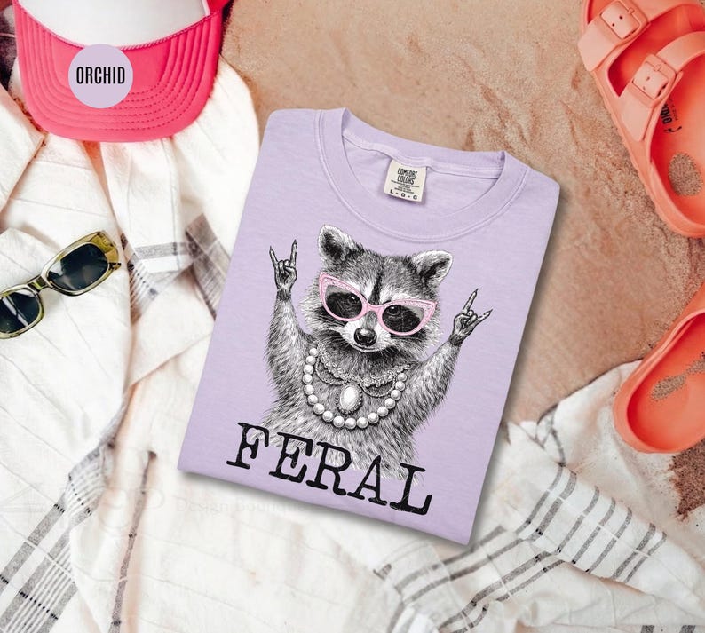 May include: A light purple t-shirt featuring a raccoon wearing pink sunglasses and a pearl necklace, with the word "FERAL" printed in black. The raccoon is making a rock and roll hand gesture. The shirt is surrounded by a pink hat, sunglasses, and sandals.