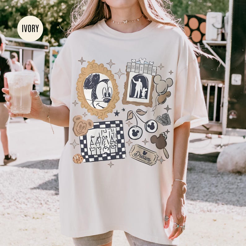 May include: Ivory-colored t-shirt featuring a collage of Disney-themed graphics. The design includes a Mickey Mouse portrait, a silhouette of a person with a dog, and various other elements like cherries and a cookie. The word "Dream" is also visible.