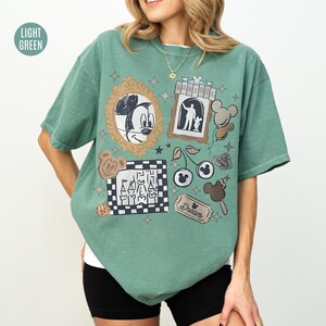 May include: Light green t-shirt with various Disney-themed graphics. The shirt features Mickey Mouse, a castle, cherries, and other cartoon elements. The word "Dream" is also visible on a ticket-shaped graphic. The shirt is a relaxed fit.