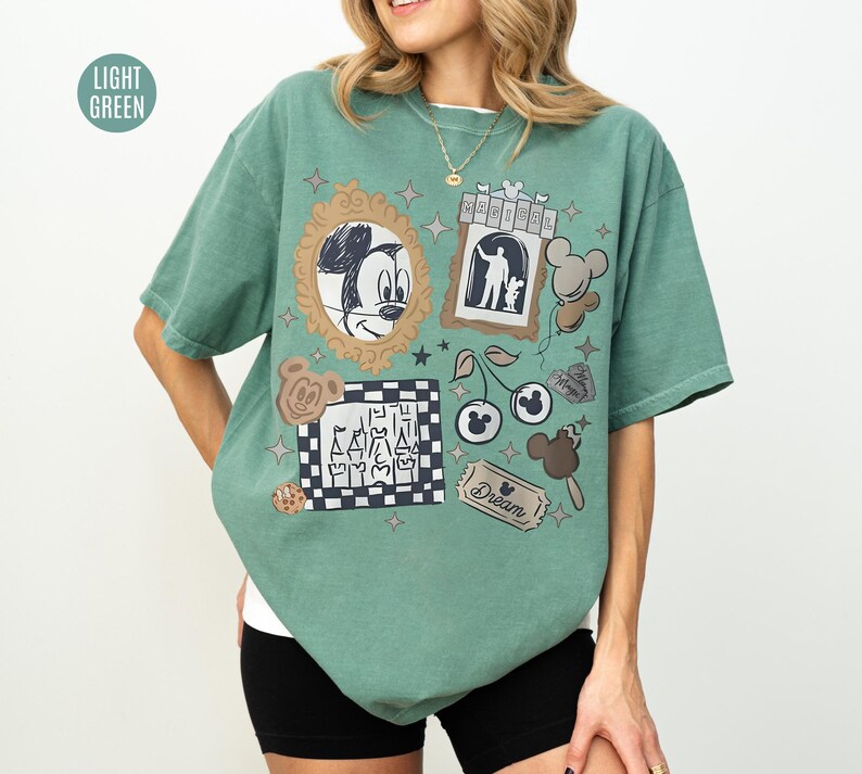 May include: Light green t-shirt with various Disney-themed graphics. The shirt features Mickey Mouse, a castle, cherries, and other cartoon elements. The word "Dream" is also visible on a ticket-shaped graphic. The shirt is a relaxed fit.
