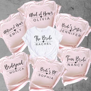 Peut inclure: Six t-shirts roses et blanches avec du texte noir et des cœurs. Les t-shirts disent "Matron d'honneur Emily", "Demoiselle d'honneur Monica", "Dame d'honneur Olivia", "La mariée Rachel", "Meilleure amie de la mariée Sophia", "Sœur de la mariée Jennifer", et "Équipe de la mariée Nancy".