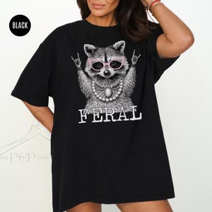 May include: Black t-shirt featuring a raccoon wearing pink glasses and a pearl necklace, with the word "FERAL" in white block letters. The raccoon is making a rock and roll hand gesture. The shirt has a relaxed fit.