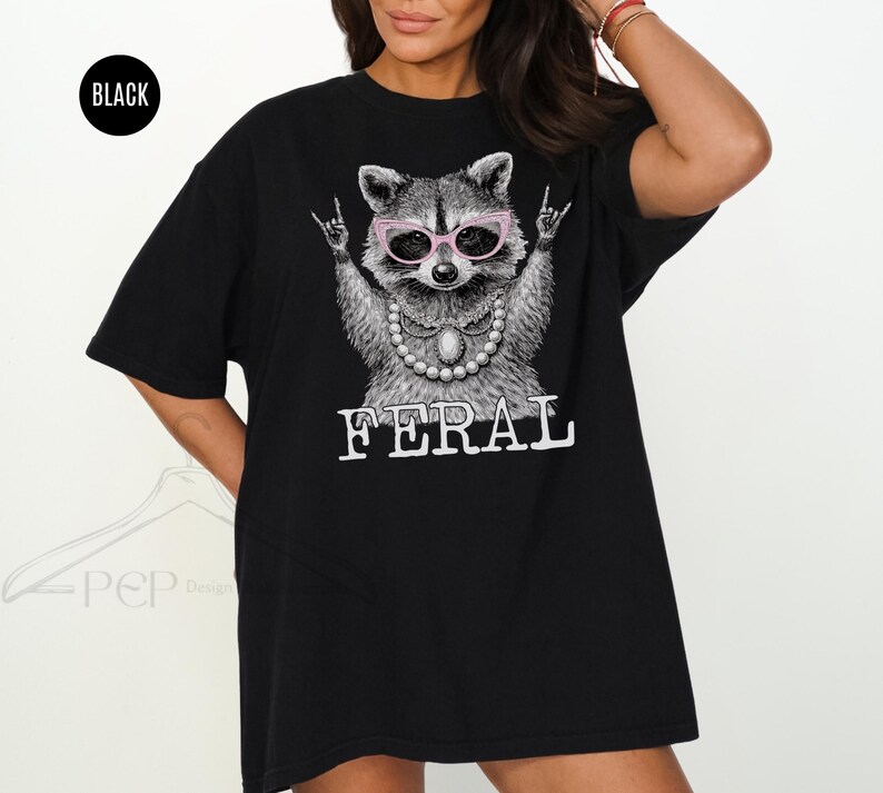 May include: Black t-shirt featuring a raccoon wearing pink glasses and a pearl necklace, with the word "FERAL" in white block letters. The raccoon is making a rock and roll hand gesture. The shirt has a relaxed fit.
