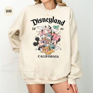 Puede incluir: Sudadera color arena con el logotipo de Disneyland, el texto "Est 1955" y "California". El dise&ntilde;o incluye a Mickey Mouse, Minnie Mouse, Donald Duck, Daisy Duck, Goofy y Pluto. La sudadera tiene cuello redondo.