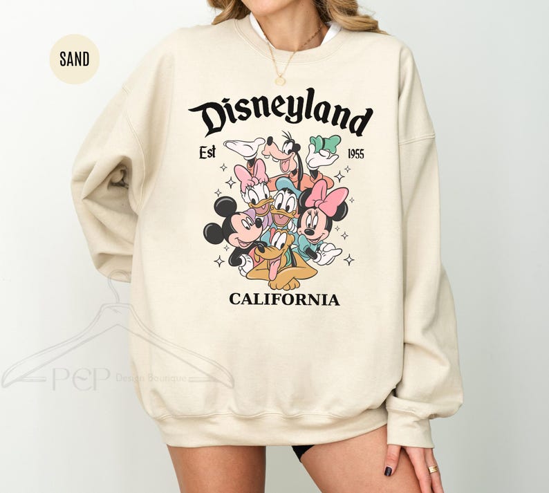 Puede incluir: Sudadera color arena con el logotipo de Disneyland, el texto "Est 1955" y "California". El dise&ntilde;o incluye a Mickey Mouse, Minnie Mouse, Donald Duck, Daisy Duck, Goofy y Pluto. La sudadera tiene cuello redondo.