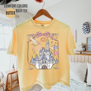 May include: Yellow Comfort Colors boxy tee shirt with a graphic of a castle, fairy, and balloons. The graphic is framed with a pink bow and floral accents. The shirt is hanging on a wooden hanger.