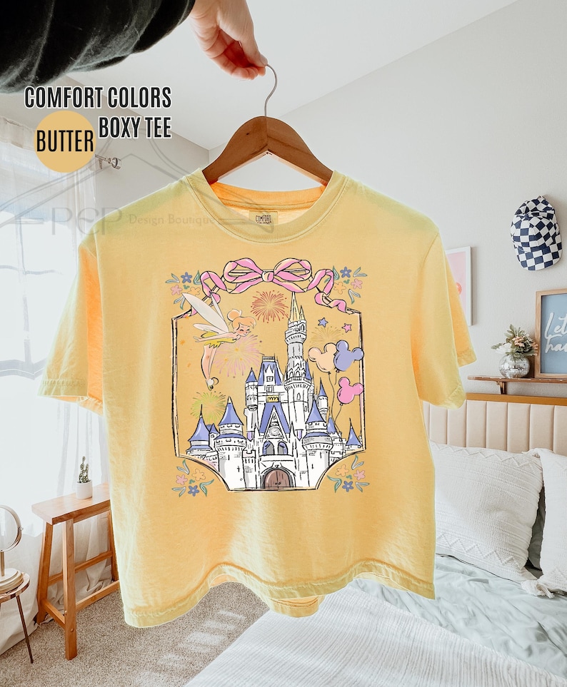 May include: Yellow Comfort Colors boxy tee shirt with a graphic of a castle, fairy, and balloons. The graphic is framed with a pink bow and floral accents. The shirt is hanging on a wooden hanger.