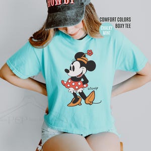 Op de afbeelding: Een lichtblauw Comfort Colors boxy tee met een Minnie Mouse-afbeelding. Het stripfiguur draagt een rode polkadot rok, een zwarte top en oranje schoenen. Het woord "Disney" staat ook op de shirt.