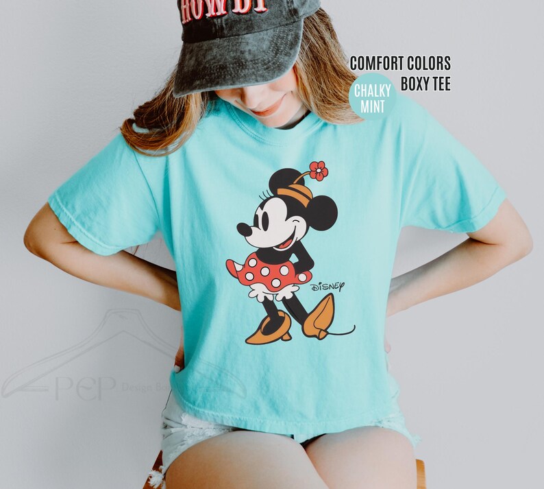 Op de afbeelding: Een lichtblauw Comfort Colors boxy tee met een Minnie Mouse-afbeelding. Het stripfiguur draagt een rode polkadot rok, een zwarte top en oranje schoenen. Het woord "Disney" staat ook op de shirt.