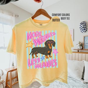May include: Butter yellow boxy tee shirt with a dachshund graphic wearing heart-shaped sunglasses. The shirt has the text "MORE WEENIES LESS MEANIES" in pink and blue. The shirt is hanging on a wooden hanger.