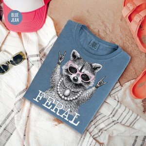 May include: Blue jean t-shirt featuring a raccoon wearing pink sunglasses and a pearl necklace, with the word "FERAL" in white. The shirt is surrounded by a pink and white hat, sunglasses, and orange sandals.