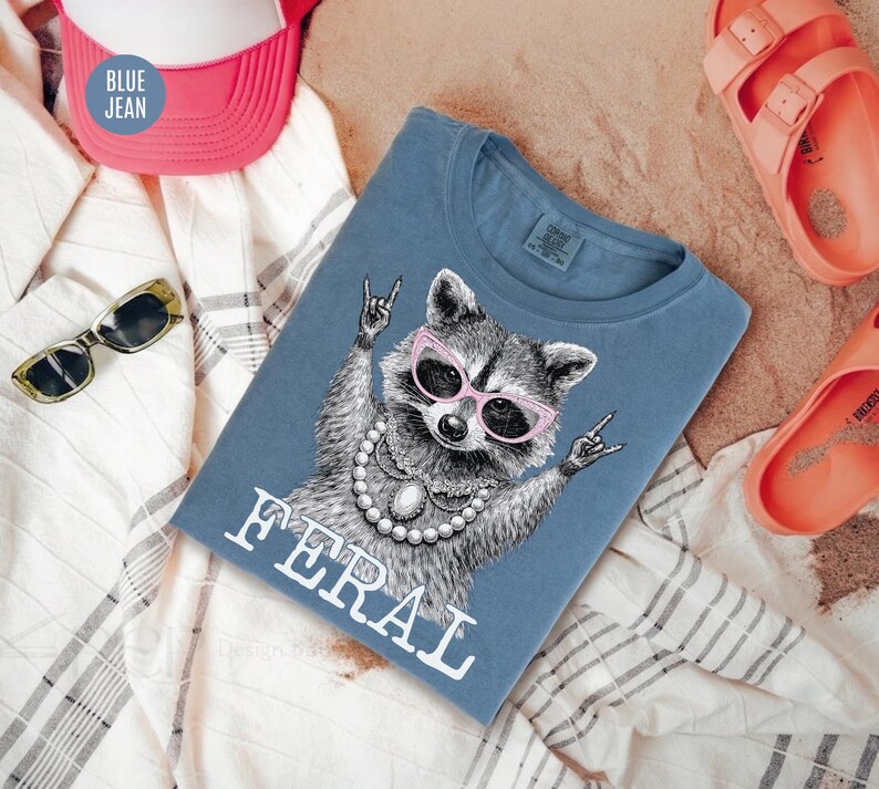 May include: Blue jean t-shirt featuring a raccoon wearing pink sunglasses and a pearl necklace, with the word "FERAL" in white. The shirt is surrounded by a pink and white hat, sunglasses, and orange sandals.