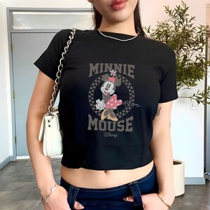 K&ouml;nnte beinhalten: Schwarzes Crop-Top mit Minnie Mouse-Grafik. Das Design zeigt die Worte "MINNIE MOUSE" in einer Vintage-Schrift, mit Minnie Mouse in einem gepunkteten Kleid. Das Disney-Logo ist ebenfalls sichtbar.